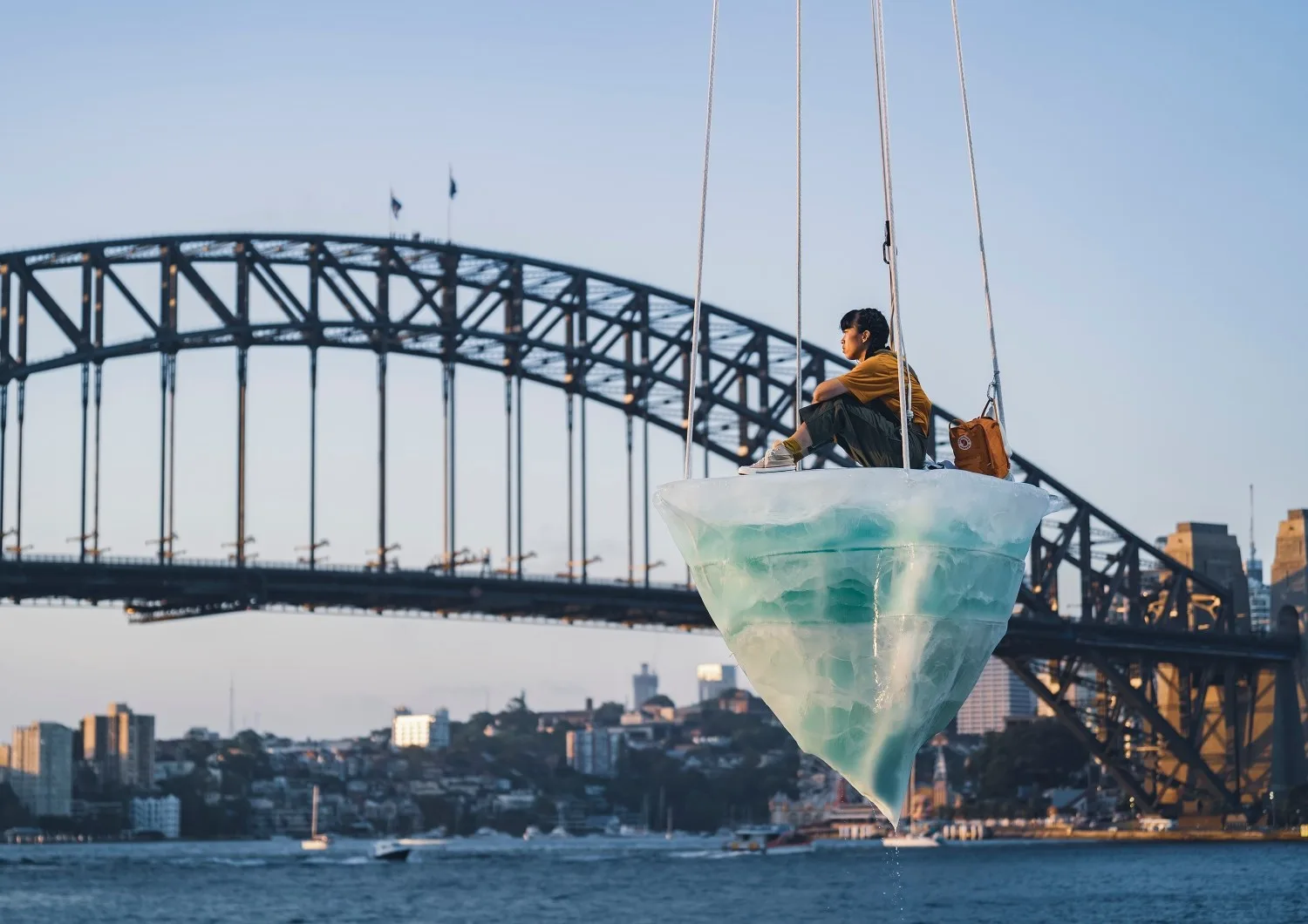 Person sitting on an iceberg sculpture suspended near Sydney Harbour Bridge with city skyline in the background.