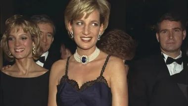 A woman in a dark dress and pearl necklace with a sapphire pendant smiles at a formal event, surrounded by people in suits.