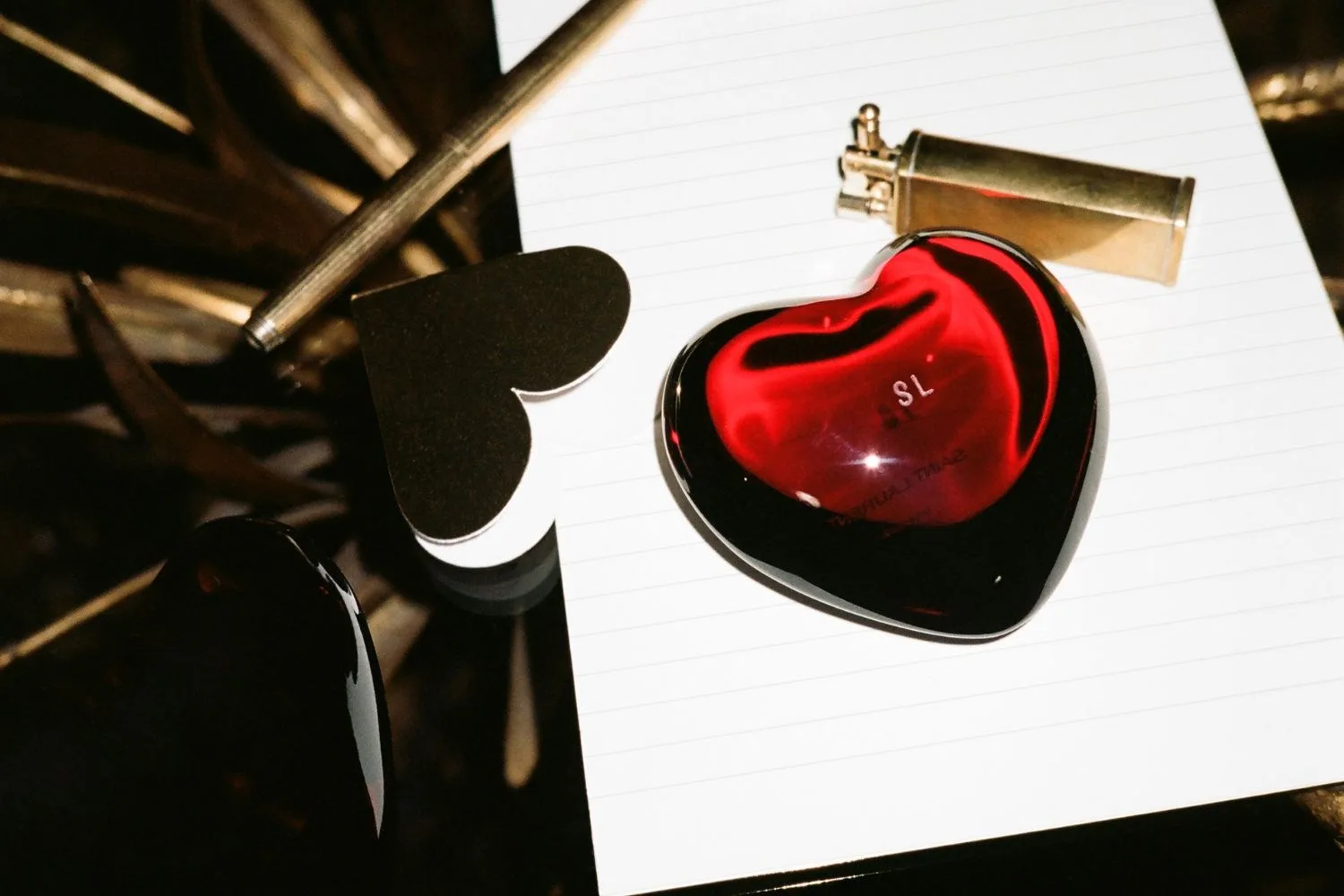 Heart-shaped red glass object on lined paper with pen, black heart, and gold lighter.