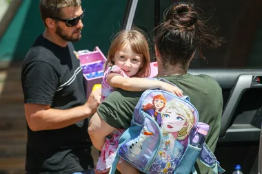 Man carrying a box, woman holding a child with a "Frozen" themed backpack featuring Elsa, Anna, and Olaf.