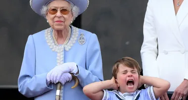 Elderly woman in blue beside a young boy covering his ears, appearing to shout.