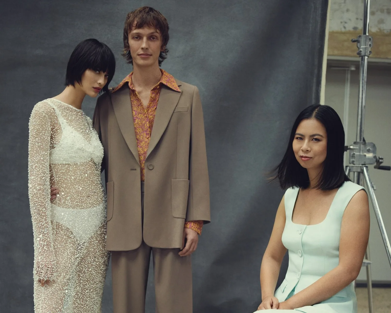 Three people posing; one in a sheer dress, another in a suit, and one seated in a light-colored dress, against a studio backdrop.