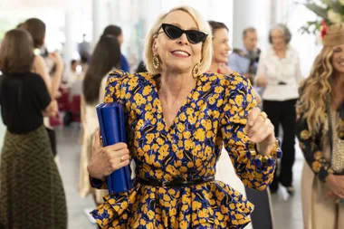 Woman in a blue and yellow floral dress, black sunglasses, holding a blue clutch at a social event.