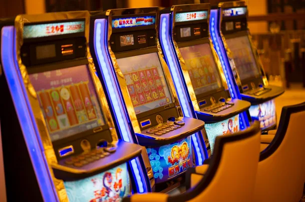 Row of brightly lit slot machines in a casino setting with chairs in front.