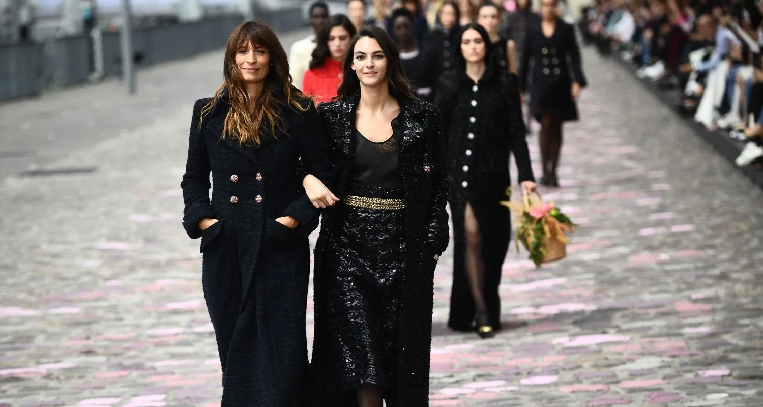 Models walk down a cobblestone runway in elegant black coats during a fashion show.