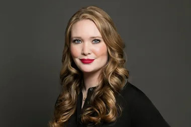A woman with long, curly blonde hair and red lipstick smiles in a professional headshot against a dark gray background.