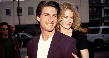 A man in a dark jacket and white shirt smiling in front of a woman with curly blonde hair wearing a black top, on a city street.