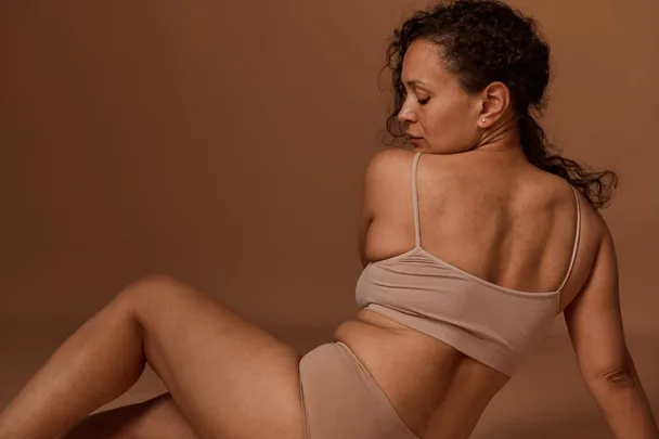 Woman with curly hair in beige clothing poses against a brown background.