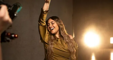 A woman with long hair smiles joyfully while raising an arm in a warmly lit room.