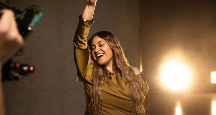 A woman with long hair smiles joyfully while raising an arm in a warmly lit room.