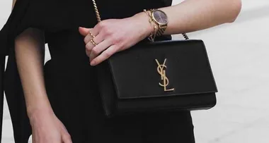 A person wearing a black outfit holds a black YSL handbag and wears a gold watch and rings.