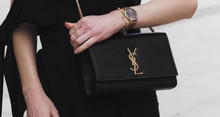 A person wearing a black outfit holds a black YSL handbag and wears a gold watch and rings.