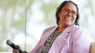 Smiling woman with red glasses holding a microphone, wearing checkered dress and lavender blazer, seated indoors.