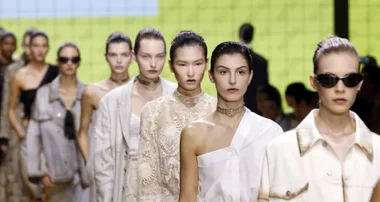 Models walking down a runway in various outfits at a fashion show, with a yellow background behind them.