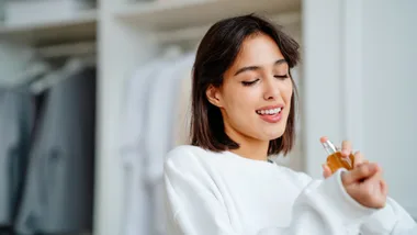 Woman in a white sweater applying perfume on her wrist, with a light background of hanging clothes.