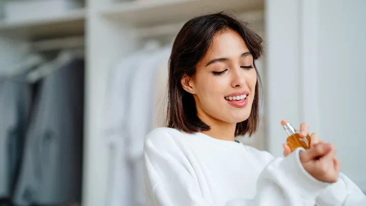 Woman in a white sweater applying perfume on her wrist, with a light background of hanging clothes.