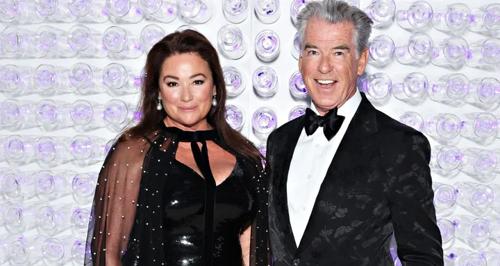 A man in a tuxedo and a woman in a black dress stand in front of a wall with purple lights.