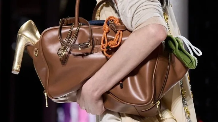 Woman holding a large brown leather handbag with gold chain and high heel detail.