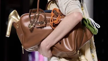Woman holding a large brown leather handbag with gold chain and high heel detail.