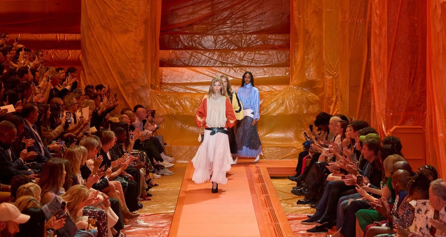 Three models walk down an orange runway lined with an audience at a fashion show.
