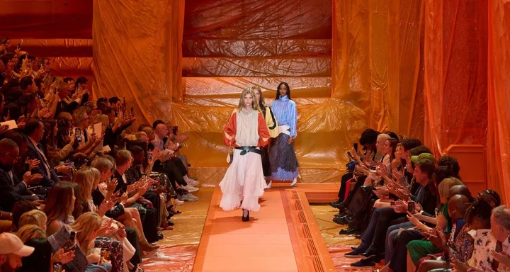 Three models walk down an orange runway lined with an audience at a fashion show.