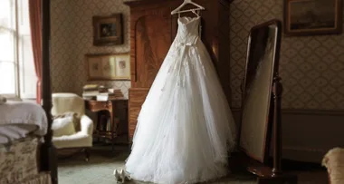 Wedding dress on hanger in vintage room beside a mirror, with shoes on the floor and a window to the left.