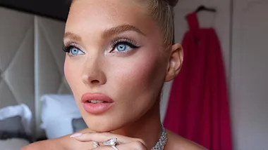 A person with blue eyes wearing a diamond necklace, pink lip makeup, and a red dress in the background.
