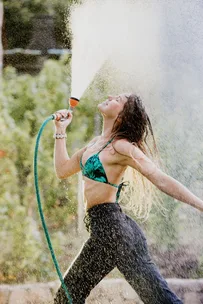 A woman in a bikini top sprays water from a hose, smiling under the sun with a lush green background.