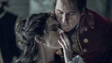 A woman in a white dress is gently shushing a man in ornate, red clothing, creating a dramatic and intimate scene.