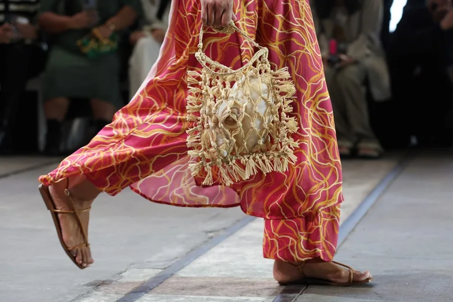 A model in sandals and a pink patterned dress walks, holding a fringed woven bag on a runway.
