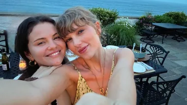 Two women smiling and taking a selfie at an outdoor dining table with a scenic ocean view in the background.