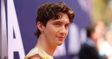 Young man with wavy hair smiling at a red carpet event, standing in front of a blurred blue and white backdrop.