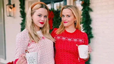 Two women in festive sweaters hold mugs with candy canes, standing outdoors with holiday greenery in the background.