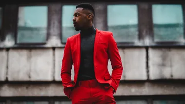 Man in a red suit and black turtleneck stands confidently in front of a modern building background.