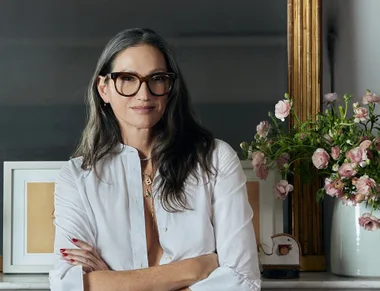 A person with long hair and glasses, wearing a white shirt, stands by a mirror with pink flowers in a vase.