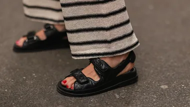 Close-up of a person wearing black sandals and striped pants, showing red-painted toenails on a sidewalk.