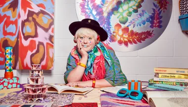 Woman in colorful attire, hat and face paint, sits thoughtfully surrounded by vibrant art and design books.