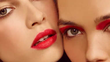 Two women pose, one is wearing a matte red lipstick while the other has a bold wash of red eyeshadow on her eyes.