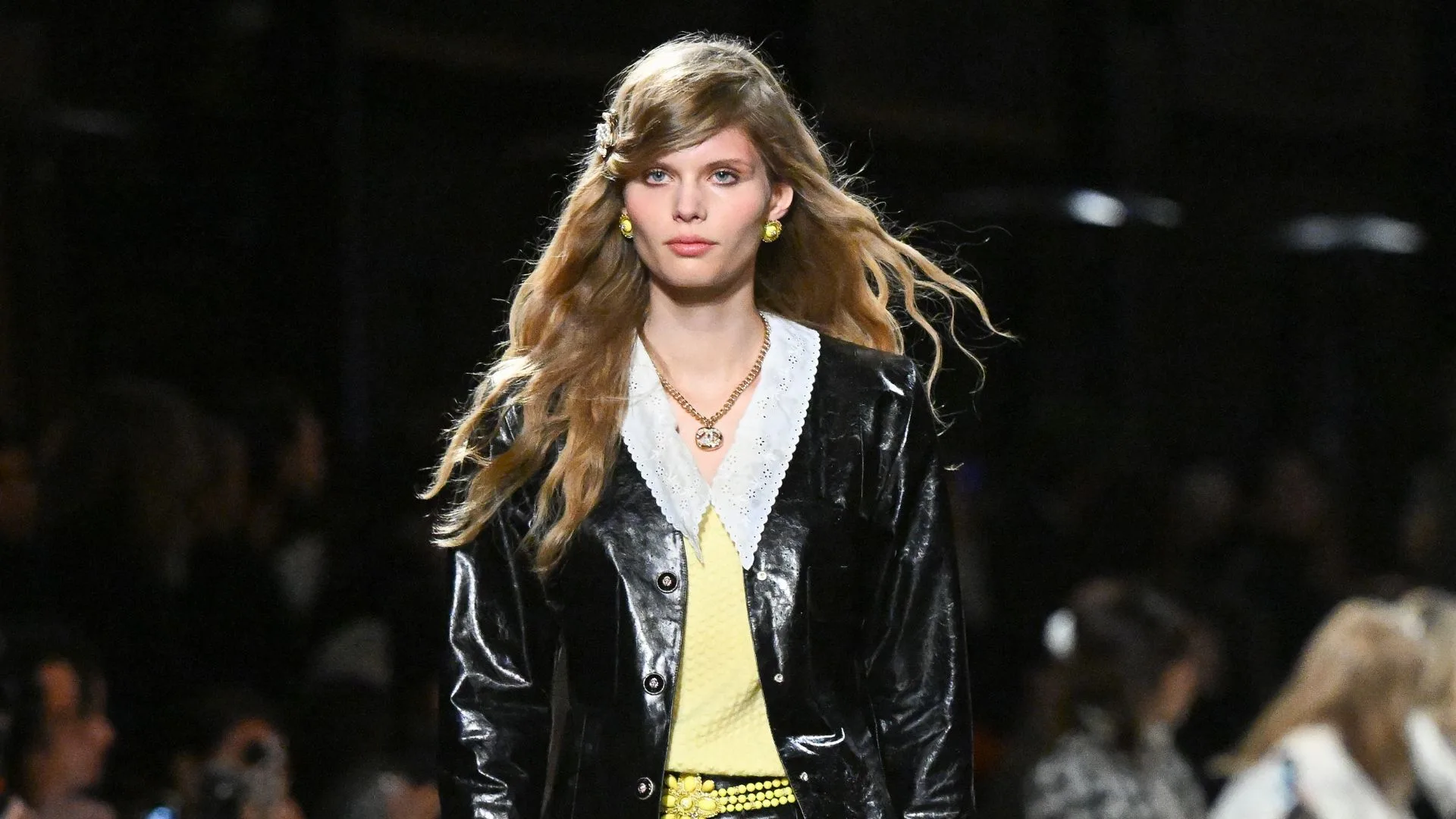 A fashion model walks the runway in a shiny black coat with a white lace collar and long wavy hair.