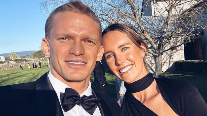 cody simpson and emma mckeon pose against a blue sky. Cody wears a black suit and bowtie, emma wears a black dress