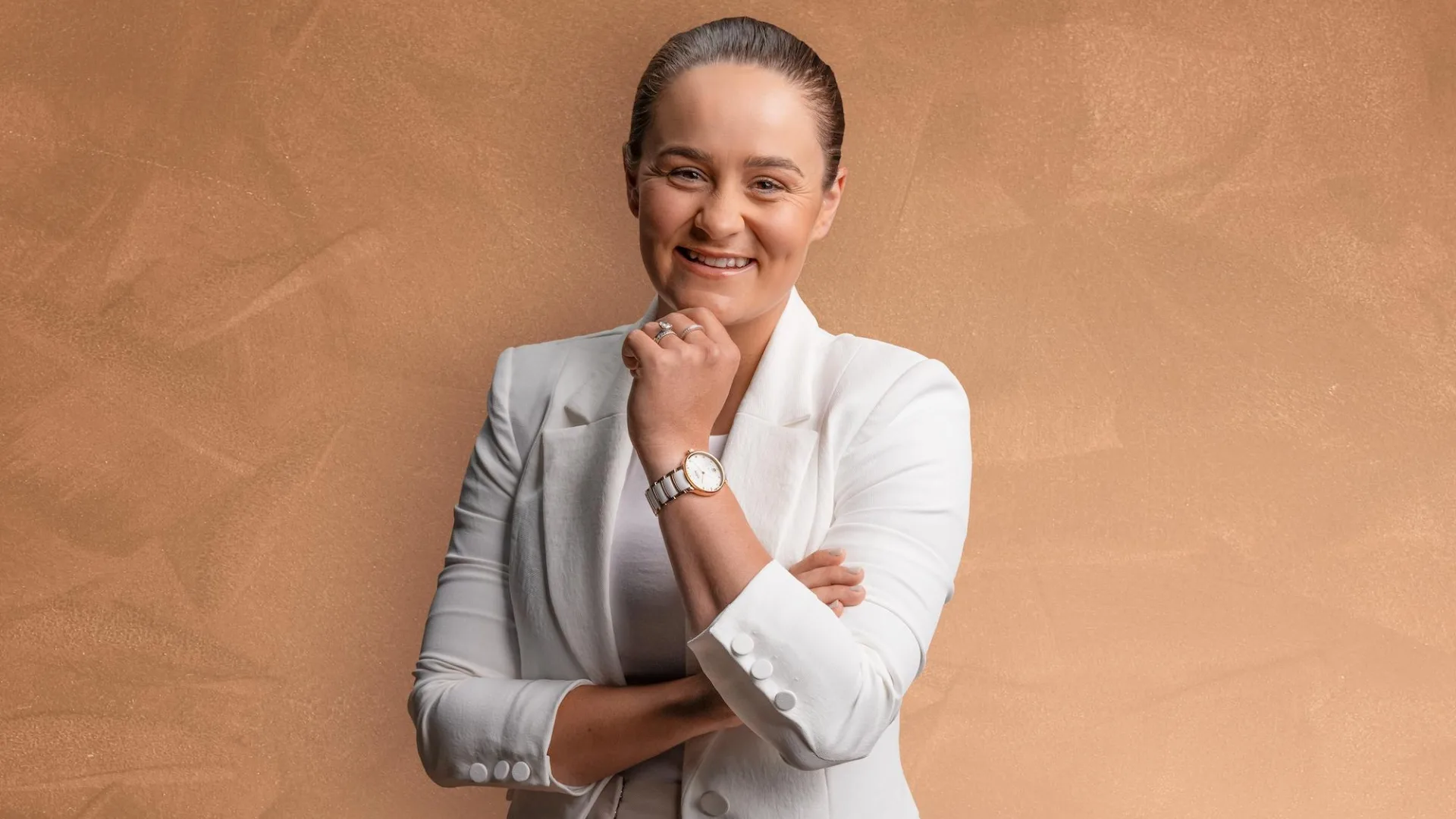 Person in a white suit smiling with arms crossed against a light brown background.
