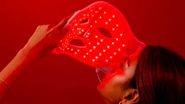 A woman holds a glowing red LED face mask in a dark red-lit room.