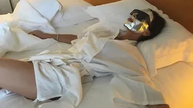 Person lying on a bed in a white robe, wearing sunglasses and a metallic facial mask.