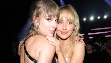 Two women with blonde hair pose closely together, smiling at the camera under bright stage lights.