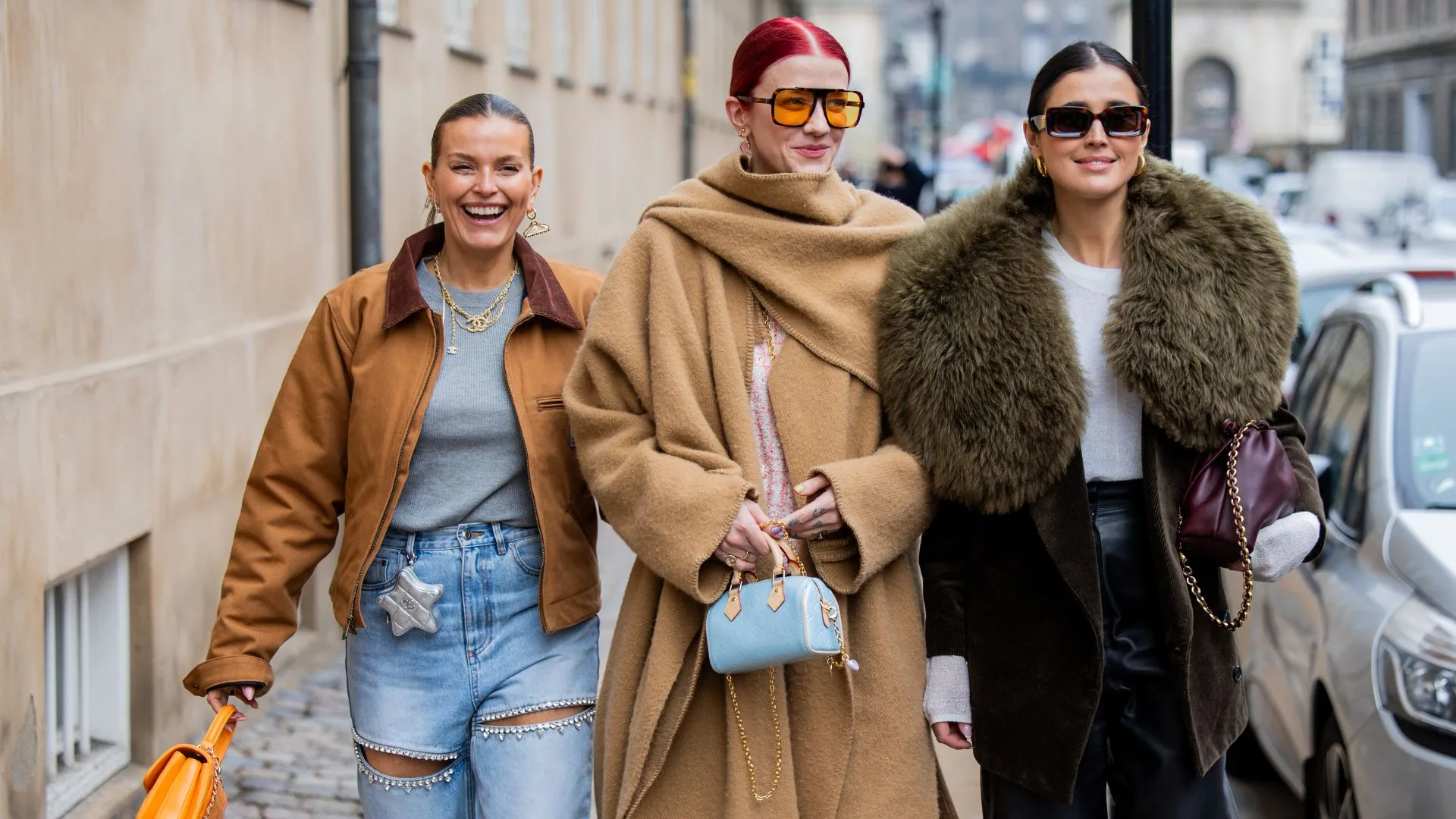 Three women in stylish winter outfits walking on a city street, smiling and carrying handbags.