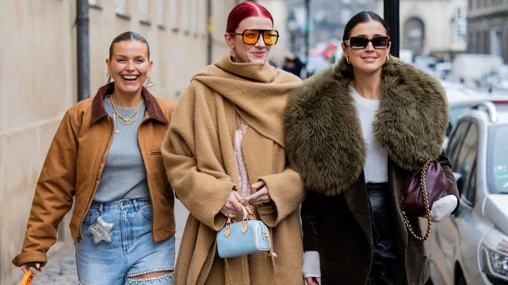 Three women in stylish winter outfits walking on a city street, smiling and carrying handbags.