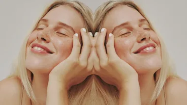 A woman with closed eyes, smiling, mirrored; blonde hair and freckled skin.