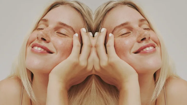 A woman with closed eyes, smiling, mirrored; blonde hair and freckled skin.