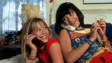 Two girls on a bed are smiling while talking on phones, one wearing a red top and the other in a floral dress.