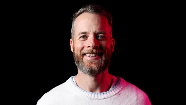 Smiling man with a beard and mustache, wearing a white sweater with red lighting accent on a black background.